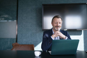 Lächelnder Geschäftsmann mit verschränkten Händen vor einem Laptop im Büro - FKF04352