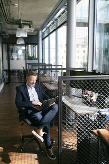Männlicher Berufstätiger, der einen Laptop benutzt, während er in einem Büroraum sitzt - FKF04365