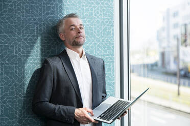 Mature businessman with laptop leaning on wall by window at office - FKF04367