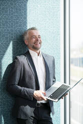 Happy businessman holding laptop in front of wall at office - FKF04368