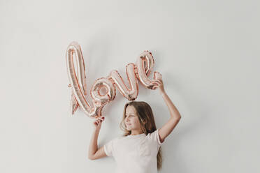 Smiling girl holding love balloon while standing in front of wall - EBBF03737