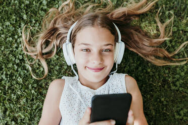 Smiling girl wearing headphones lying on grass - EBBF03749