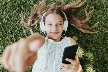 Smiling girl wearing headphones pointing while lying on grass - EBBF03750