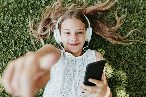 Lächelndes Mädchen mit Kopfhörern, das im Gras liegend auf etwas zeigt - EBBF03750