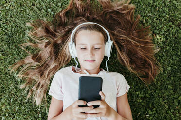 Girl wearing headphones listening music while lying on grass - EBBF03752