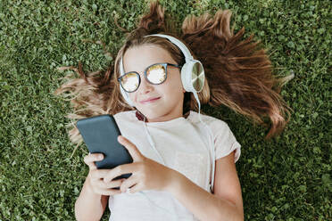 Cute girl wearing sunglasses listening music while lying on grass - EBBF03753