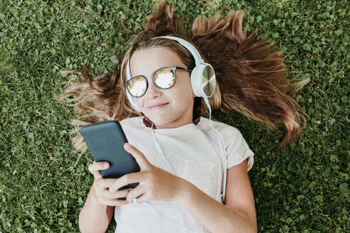 Nettes Mädchen mit Sonnenbrille hört Musik, während sie im Gras liegt - EBBF03753