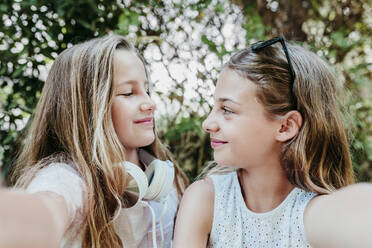 Smiling girls looking at each other while taking selfie in garden - EBBF03762