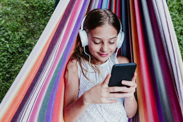 Smiling girl wearing headphones using mobile phone while resting in hammock - EBBF03769