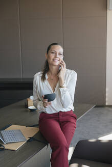 Lächelnde Unternehmerin, die eine Kaffeetasse hält und mit ihrem Smartphone spricht, während sie am Schreibtisch im Büro sitzt - JRVF00790