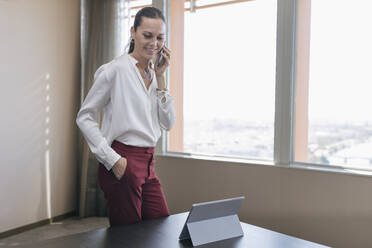 Lächelnde reife Geschäftsfrau, die auf einem Smartphone spricht, während sie am Schreibtisch im Büro steht - JRVF00795
