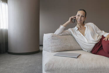 Lächelnde Unternehmerin, die auf dem Sofa sitzend mit ihrem Smartphone telefoniert und dabei wegschaut - JRVF00826