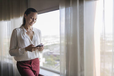 Weibliche Berufstätige, die mit ihrem Mobiltelefon SMS schreiben, während sie im Büro am Fenster stehen - JRVF00830