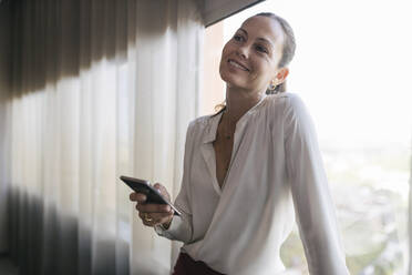 Lächelnde Geschäftsfrau stehend mit Smartphone am Fenster im Büro - JRVF00831