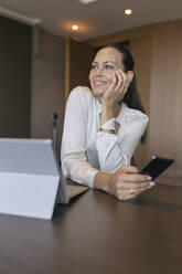 Smiling thoughtful female professional holding smart phone sitting at desk in office - JRVF00836