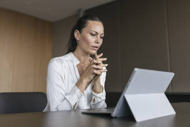 Female entrepreneur with hands clasped looking at digital tablet while working in office - JRVF00838