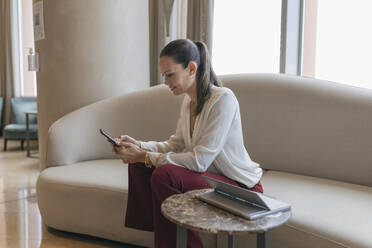 Berufstätige Frau benutzt ihr Smartphone, während sie auf dem Sofa im Büro sitzt - JRVF00842