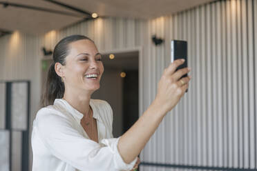 Fröhliche reife Geschäftsfrau bei einem Videoanruf über ein Smartphone in der Lobby - JRVF00849