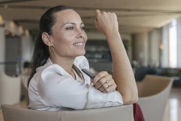 Smiling businesswoman looking away while sitting on chair in lobby - JRVF00861