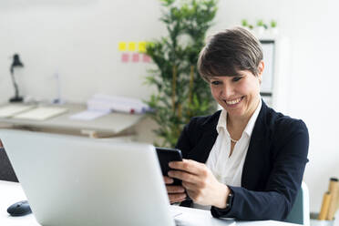 Fröhliche weibliche professionelle Textnachrichten über ein Smartphone, während sie am Schreibtisch im Büro sitzt - GIOF12681