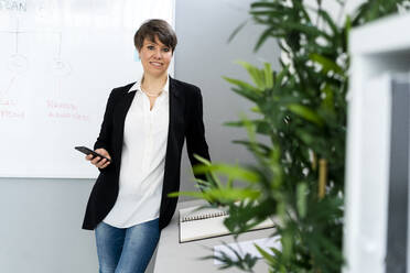 Lächelnde Geschäftsfrau mit Smartphone im Büro stehend - GIOF12683