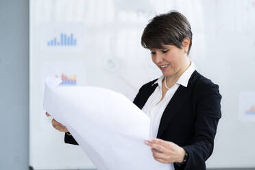 Smiling female professional analyzing paper chart while working in office - GIOF12689