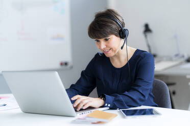 Smiling female professional wearing headset using laptop sitting at desk in office - GIOF12698