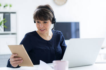 Businesswoman wearing headset sitting with laptop at desk in office - GIOF12700