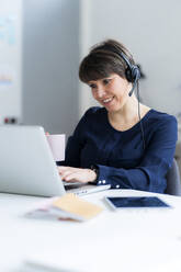 Smiling businesswoman wearing headset using laptop while working in office - GIOF12704
