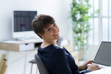 Smiling businesswoman sitting with laptop in office - GIOF12705