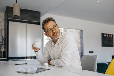 Male freelancer with coffee cup looking away while sitting in kitchen at home - JRFF05158