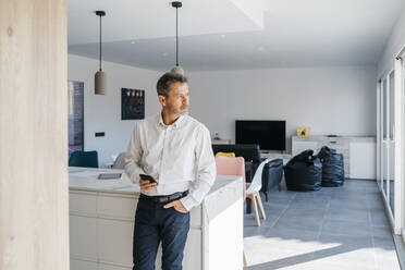 Male professional with hand in pocket holding mobile phone in front of kitchen island at home - JRFF05159