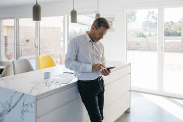 Male freelancer working on mobile phone while standing in front of kitchen island at home - JRFF05170