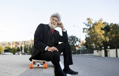 Smiling mature businessman sitting on skateboard while day dreaming - JCCMF02619