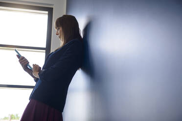 Junge Frau benutzt ein digitales Tablet, während sie sich im Büro an die Wand lehnt - BMOF00823