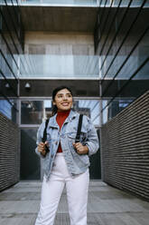 Smiling woman with backpack looking away while standing on footpath - TCEF01858