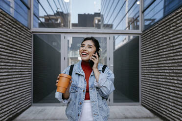 Fröhliche Frau mit Kaffeetasse, die vor einem Gebäude mit ihrem Mobiltelefon spricht - TCEF01863