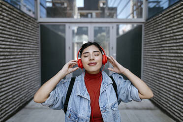 Lächelnde Frau hört Musik über Kopfhörer vor einem Gebäude - TCEF01865