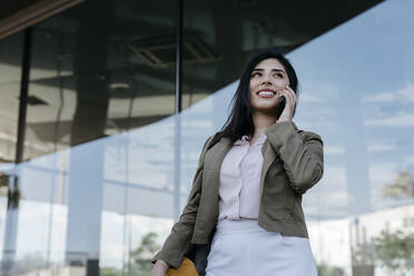 Junge Geschäftsfrau, die vor einem Gebäude mit einem Mobiltelefon spricht - TCEF01877