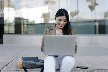 Smiling female freelancer using laptop while sitting on footpath - TCEF01878