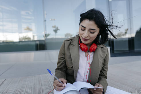 Geschäftsfrau mit Kopfhörern, die auf dem Gehweg in ein Buch schreibt - TCEF01884