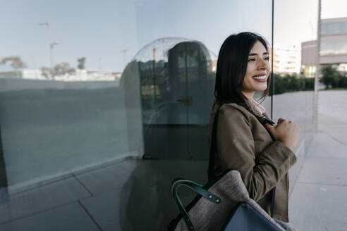 Lächelnde Geschäftsfrau mit Tasche an Glaswand gelehnt - TCEF01887