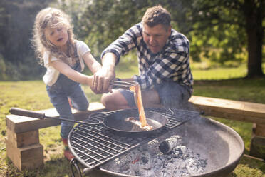 Vater und Tochter kochen Speck über einem Holzkohlegrill im Freien - CAIF30354