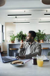 Thoughtful businessman working at laptop at breakfast in office - CAIF30412