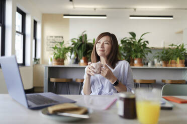 Glückliche Geschäftsfrau trinkt Kaffee am Laptop in der Bürolounge - CAIF30425