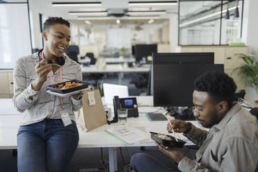 Happy business people eating takeout lunch at desk in open plan office - CAIF30446