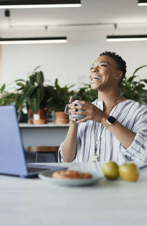 Glückliche Geschäftsfrau mit Kaffee lachend am Laptop im Büro - CAIF30486