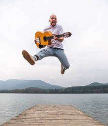 Voller Länge von bärtigen Mann in Freizeitkleidung springen mit Gitarre auf hölzernen Pier in der Nähe von See mit grünen Bergen im Hintergrund - ADSF24690