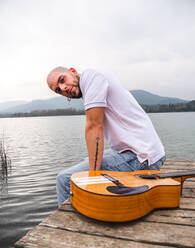 Seitenansicht Mann in Freizeitkleidung sitzt mit Gitarre auf hölzernen Pier in der Nähe von Fluss mit Bergen im Hintergrund unter bewölkten grauen Himmel in der Tageszeit - ADSF24691