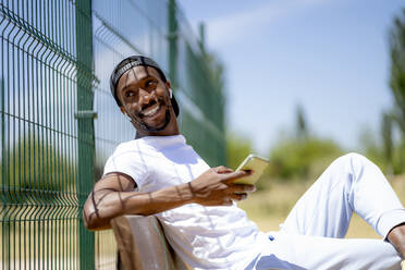 Smiling young man with mobile phone looking away at park - OCMF02118